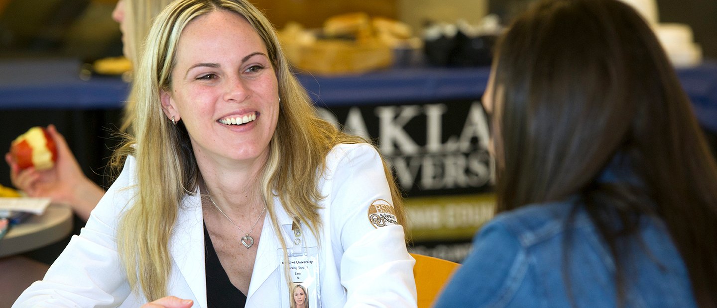 SON-19888_student-orgs.jpg woman in a white lab coat smiling, speaking to another woman in front of her